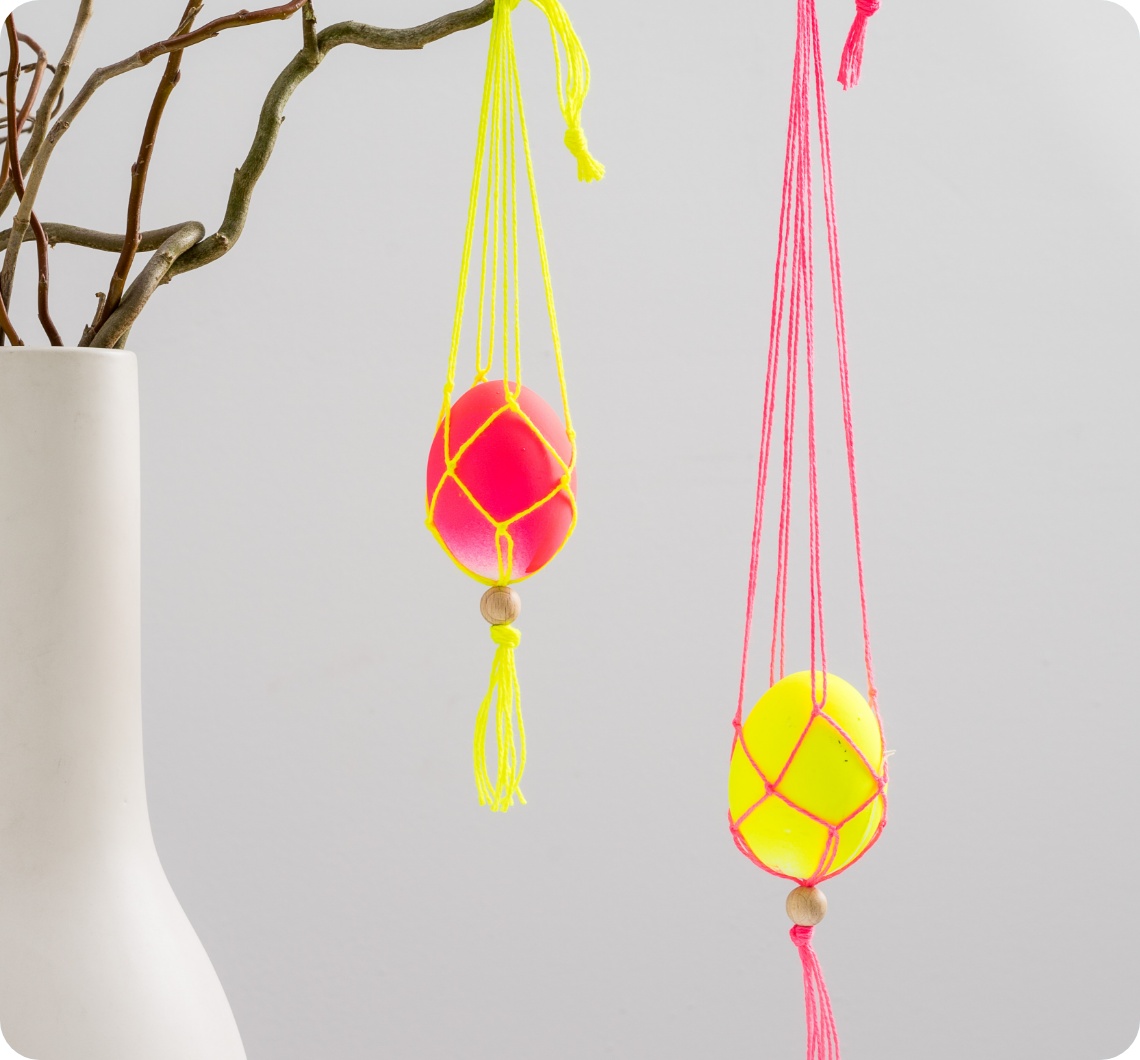 Bunte Ostereier in Makramee-Netzen aus gelben und pinken Fäden, hängend an Frühlingszweigen