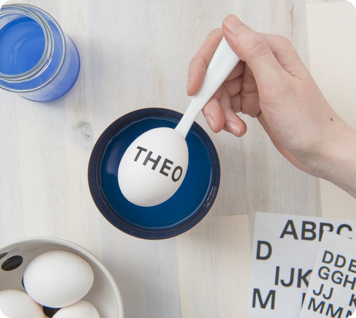 Weiße Hand taucht ein Osterei mit THEO-Schriftzug in blaue Farbe, daneben Klebebuchstaben und ein Glas mit blauer Flüssigkeit.