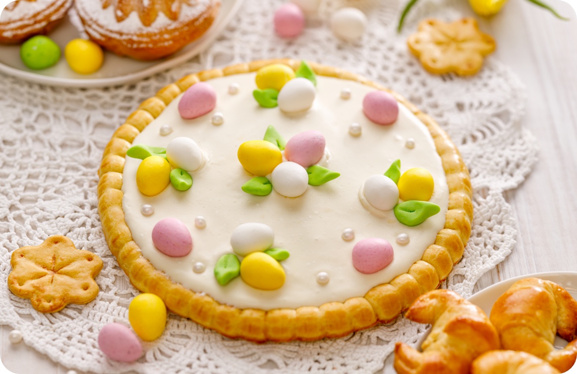 Osterkuchen mit weißem Zuckerguss, bunten Deko-Eiern und Marzipanblättern auf Häkeldecke serviert.