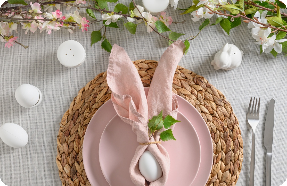 Oster-Tischdekoration mit rosafarbenem Geschirr, Ei in Serviette als Hasenohren gefaltet und blühenden Zweigen.
