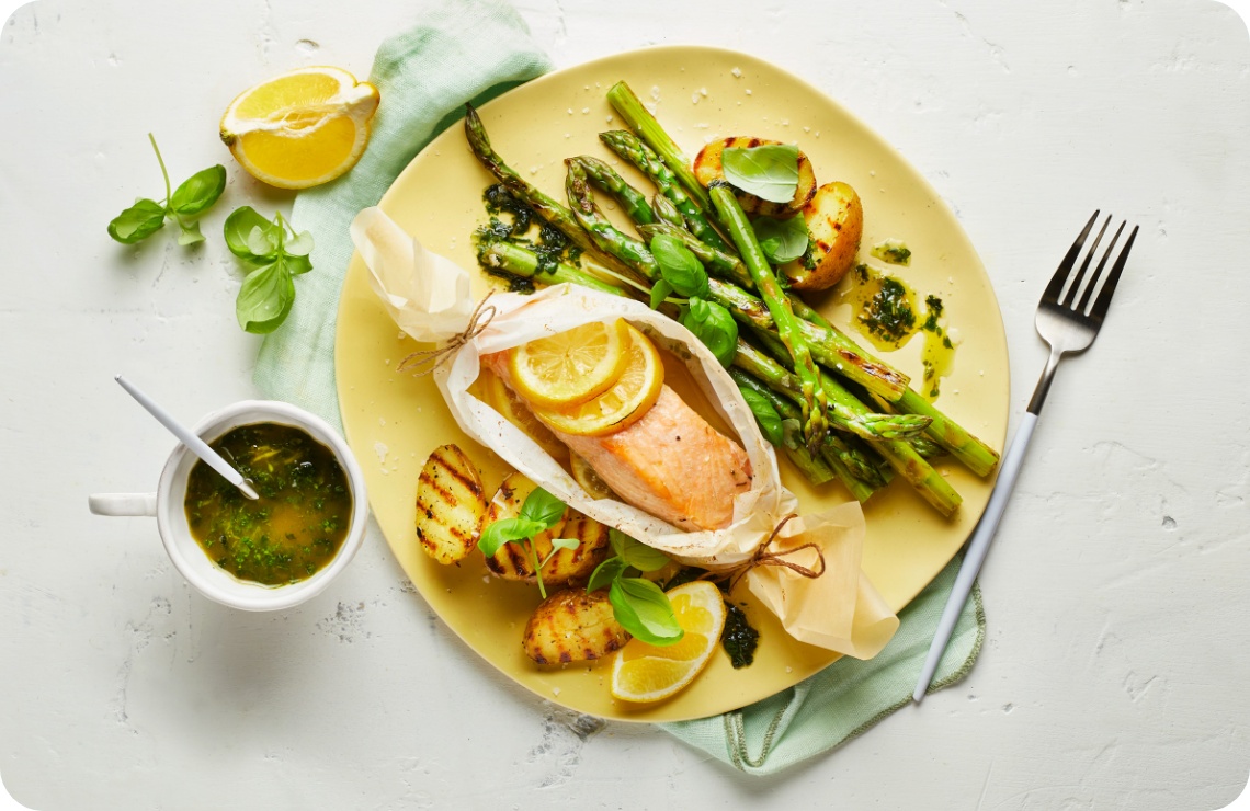 Lachs im Pergament mit Zitronenscheiben, gegrilltem Spargel und Kartoffeln auf einem gelben Teller, serviert mit Kräutersauce.