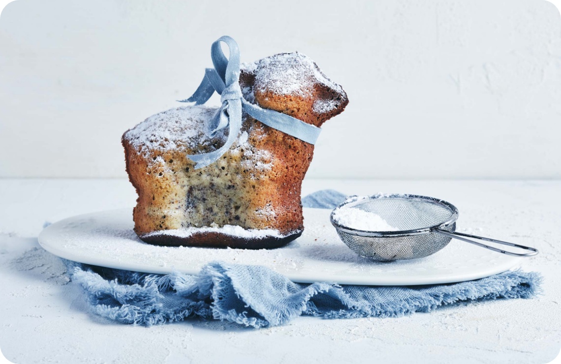 Gebäck in Lammform mit Puderzucker bestäubt, verziert mit blauer Schleife, auf weißem Teller mit Sieb und blauer Stoffserviette.