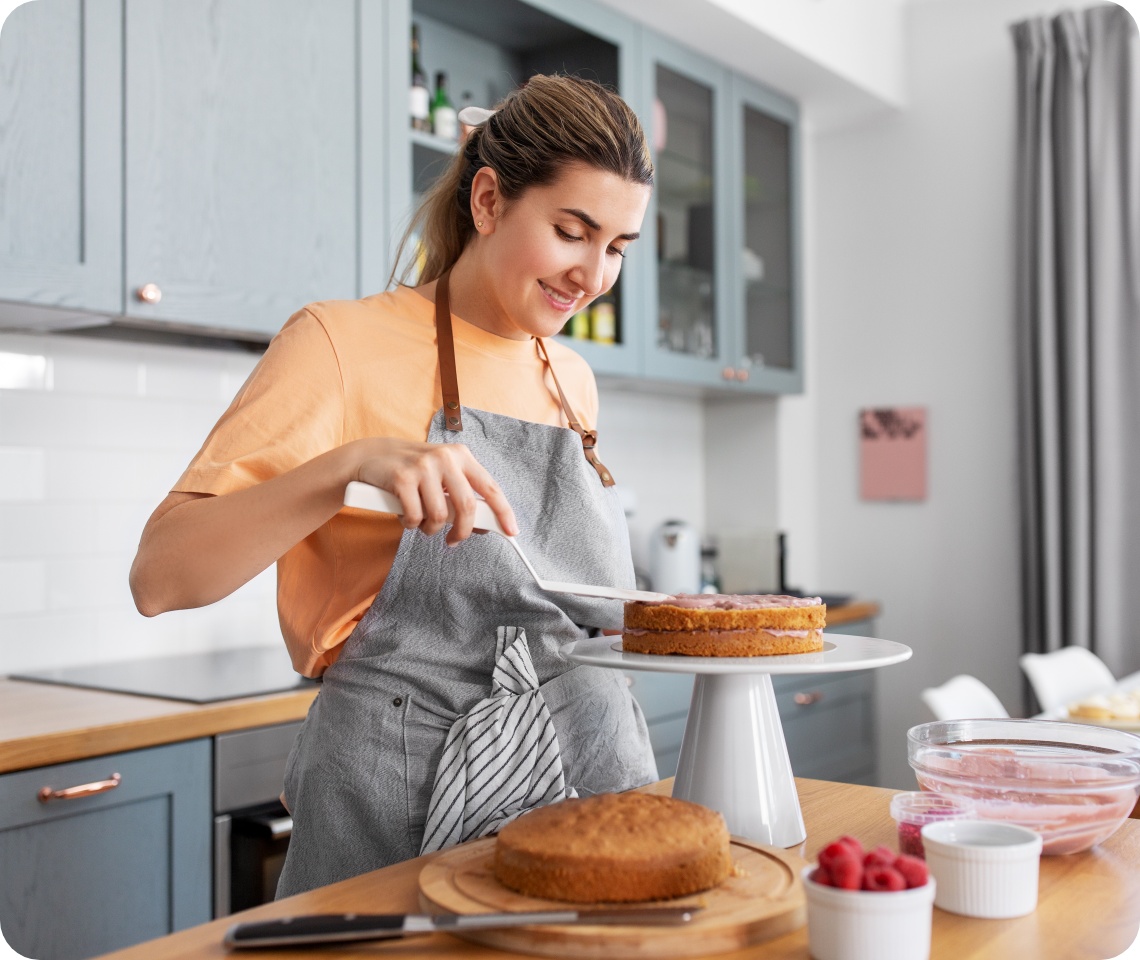 Lächelnde Frau in Schürze bestreicht einen Kuchen mit Creme in einer modernen Küche, umgeben von Backzutaten und Himbeeren.