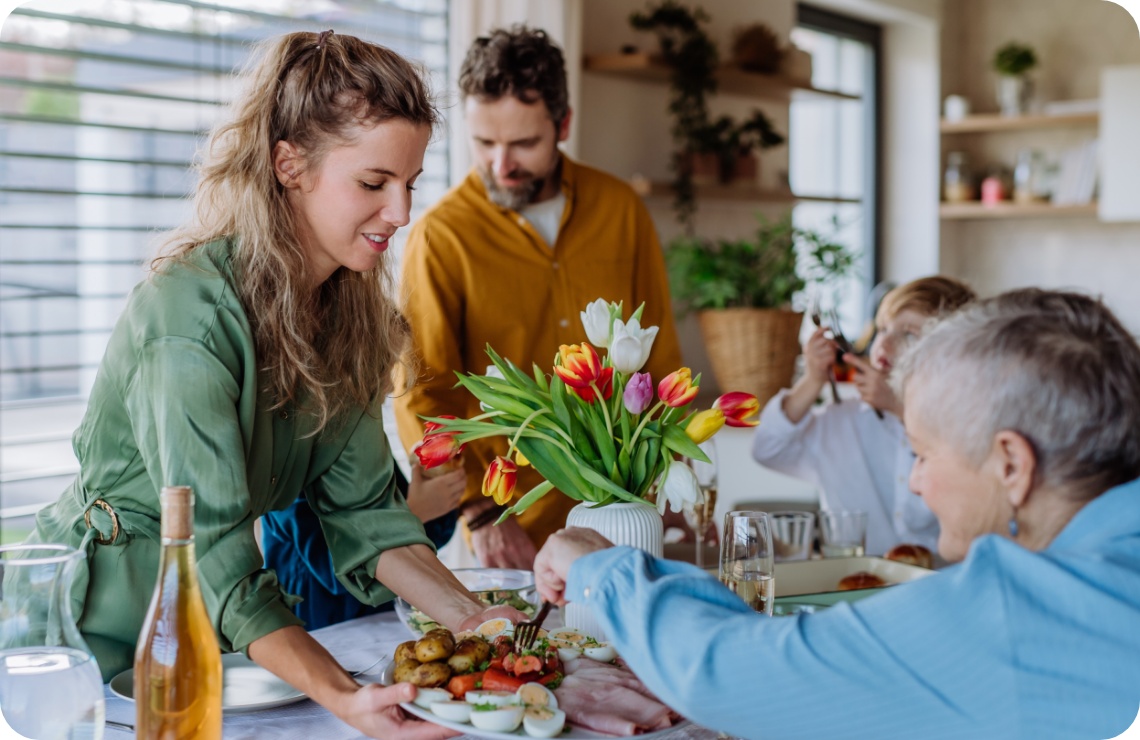 Familie genießt gemeinsames Essen an festlich gedecktem Tisch mit Tulpenstrauß, während eine Frau ein Gericht serviert.