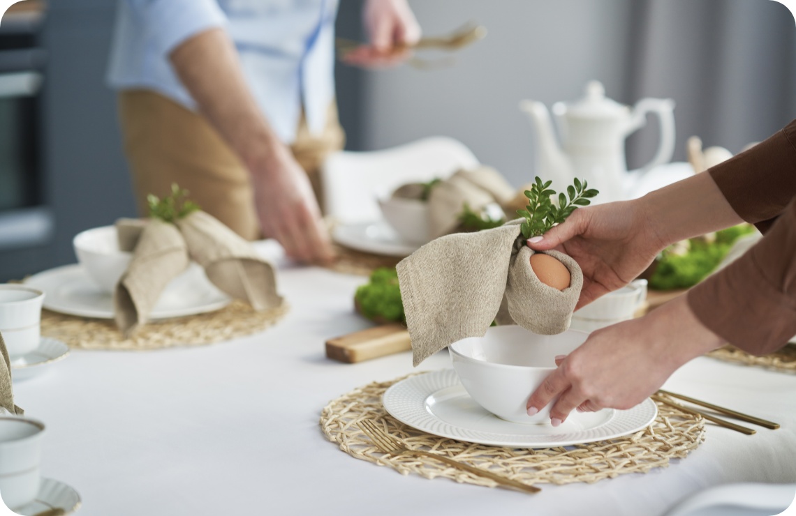 Festlich gedeckter Tisch mit handgefalteter Serviette, die ein Ei und Grünzweig umhüllt, als Osterdekoration auf einem weißen Teller.
