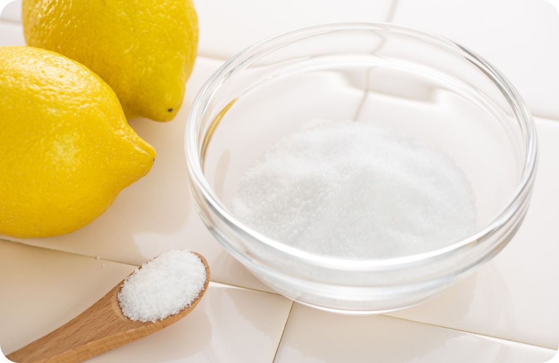 Glasschale mit Zitronensäurepulver, einem Holzlöffel und frischen Zitronen auf weißen Fliesen.