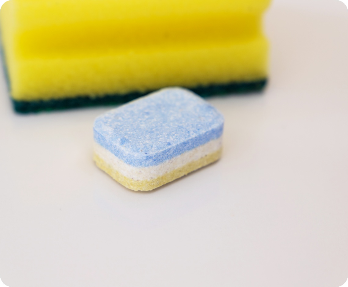 Nahaufnahme eines bunten Spülmaschinentabs vor einem gelben Schwamm auf einer weißen Oberfläche.