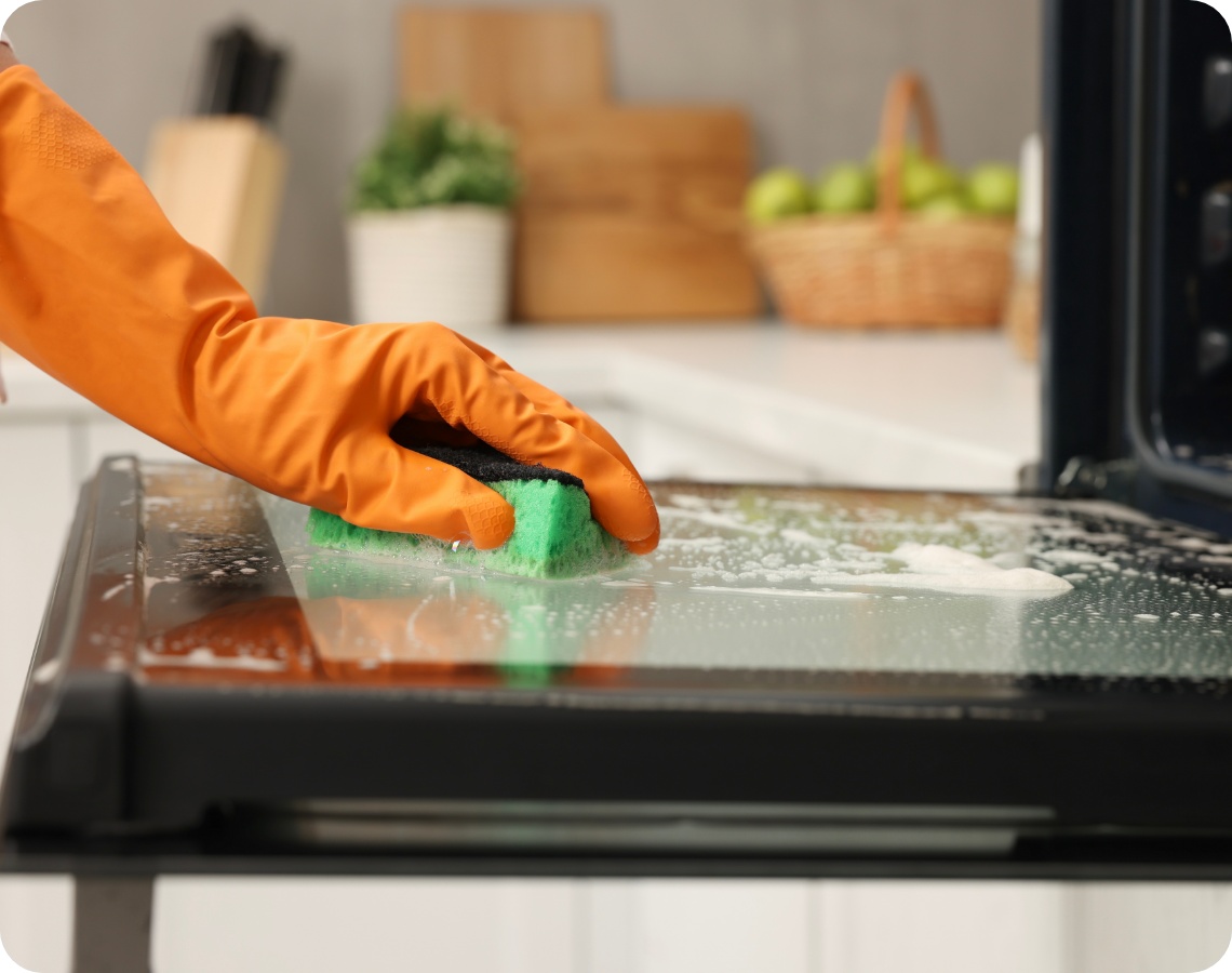 Person mit orangefarbenem Gummihandschuh reinigt die Glasinnenseite einer offenen Backofentür mit einem grünen Schwamm.
