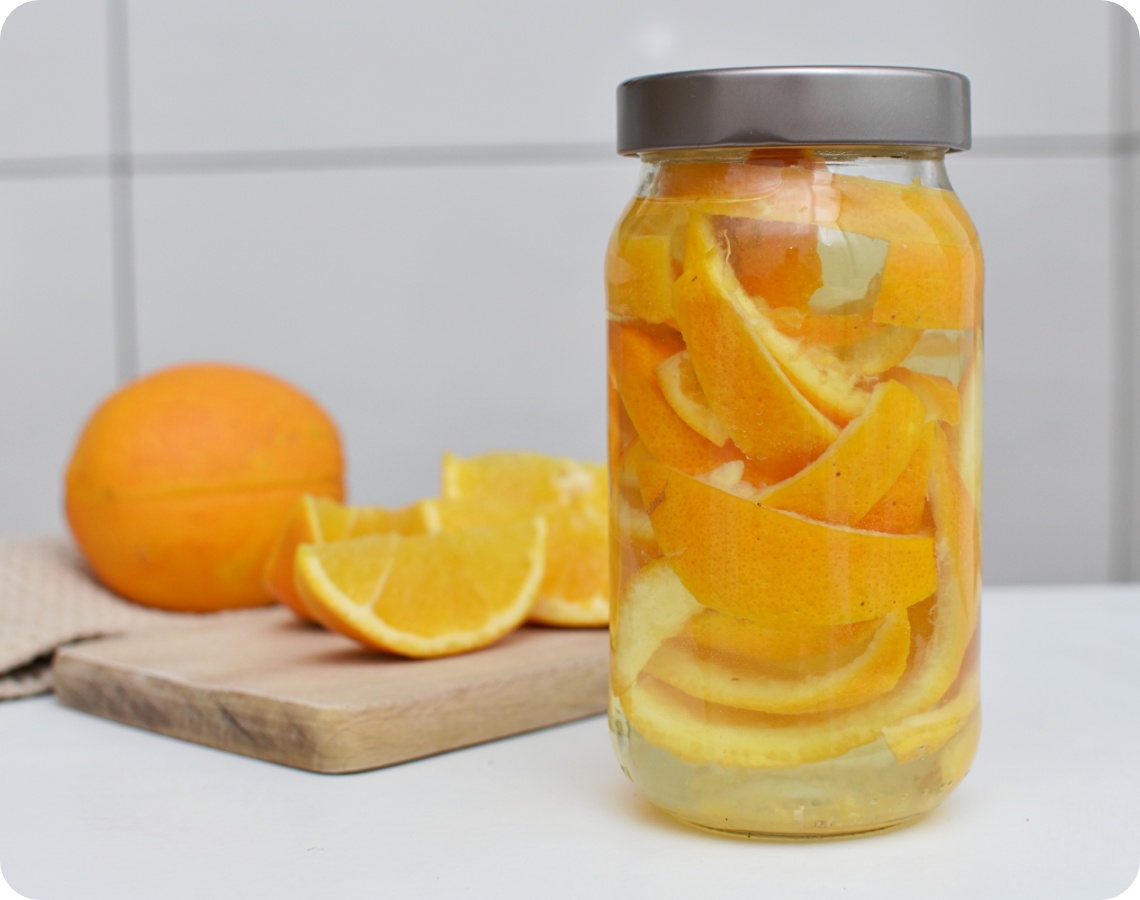 Glas mit Orangenschalen in Essig zur Herstellung eines natürlichen Reinigers, daneben frische Orangen auf einem Schneidebrett.