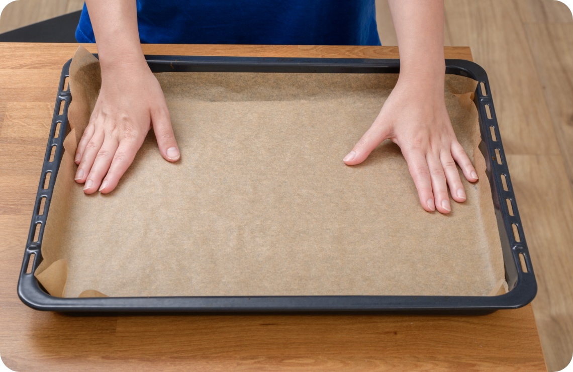 Person legt ein Stück Backpapier auf ein schwarzes Backblech, das auf einem Holztisch liegt.