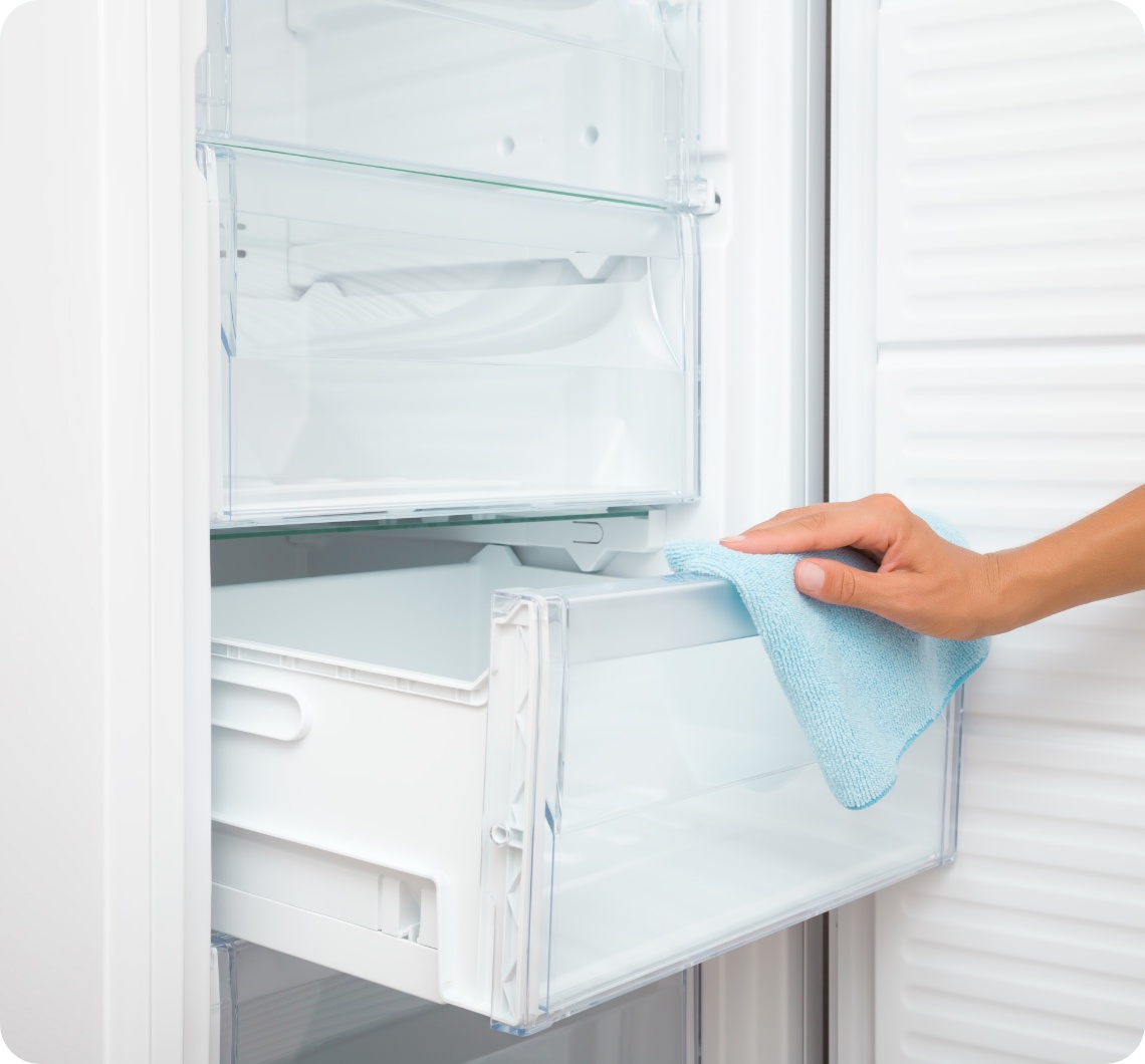 Jemand wischt mit einem blauen Mikrofasertuch die transparente Gefrierschublade eines weißen, sauberen Kühlschranks aus.