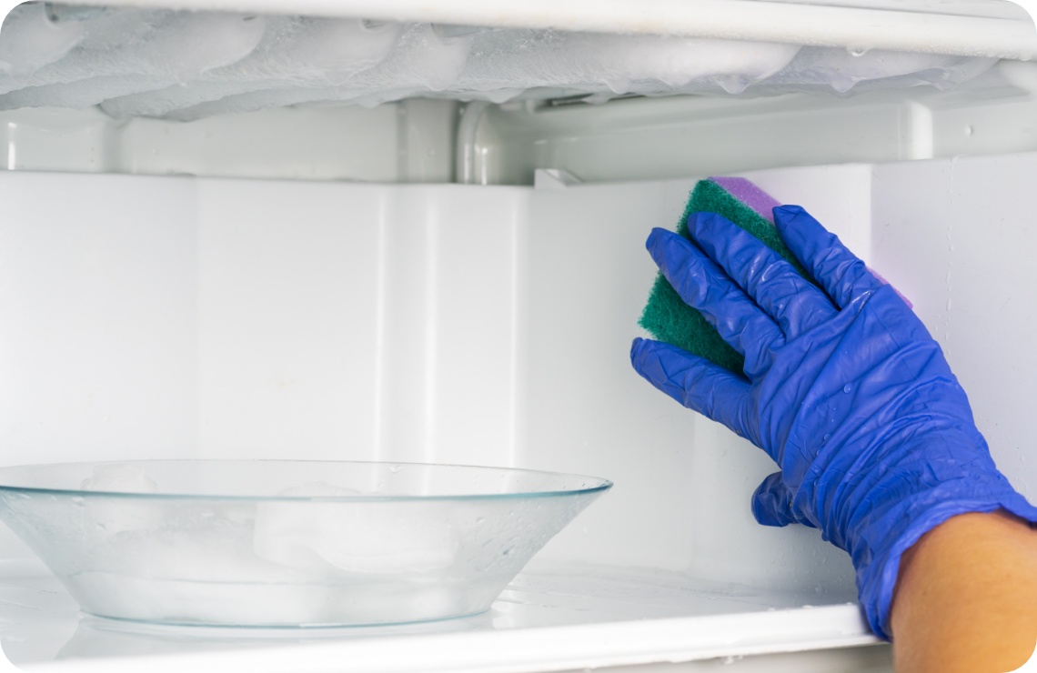 Person mit blauen Gummihandschuhen reinigt einen von innen vereisten Gefrierschrank mit einem grünen Schwamm.