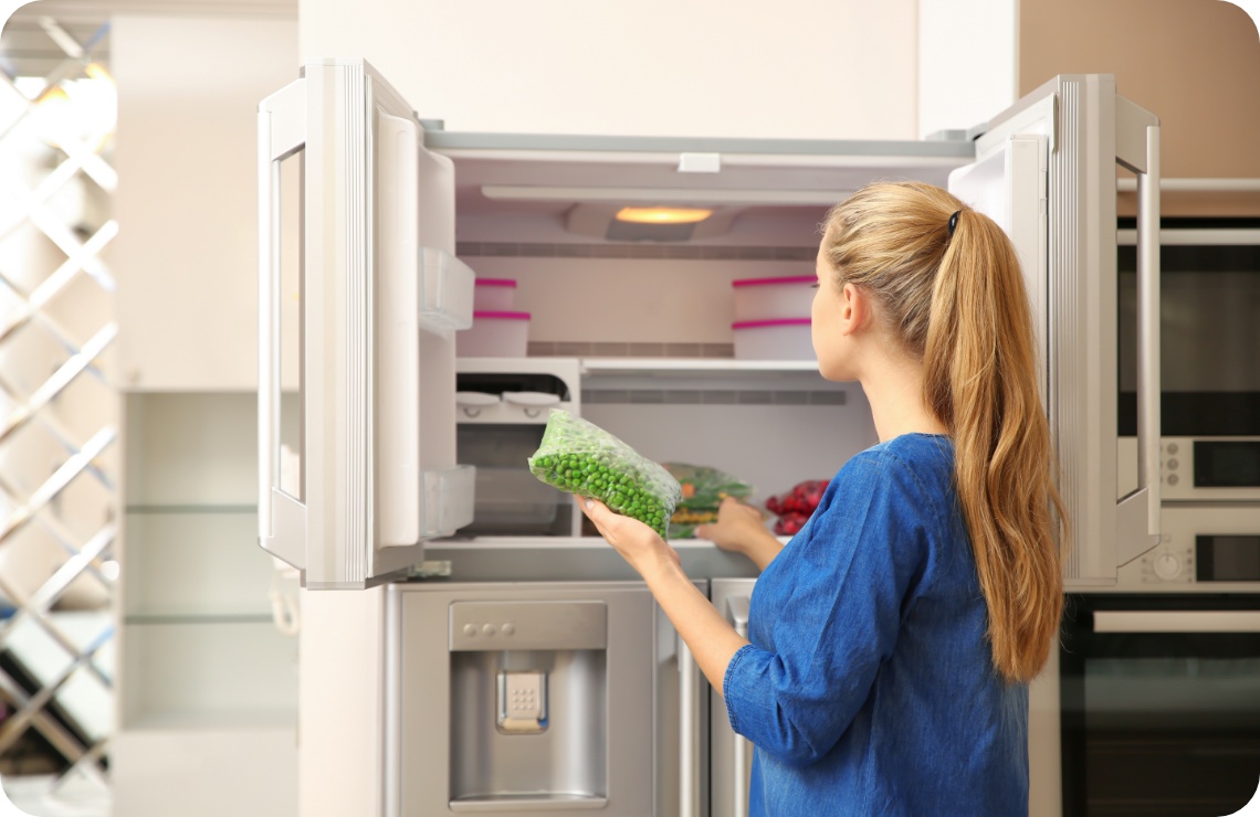 Frau legt eine Tüte gefrorene Erbsen in einen doppeltürigen Gefrierschrank in einer modernen Küche.