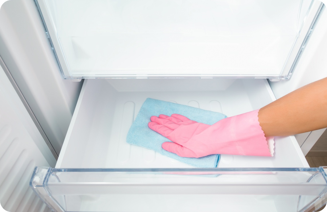 Person mit rosa Gummihandschuhen reinigt ein leeres Gefrierfrach mi einem blauen Mikrofasertuch.