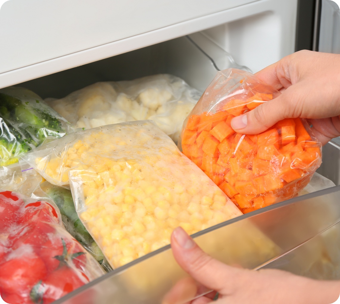 Person legt gefrorene und in einer Plastiktüte verpackte Möhrenwürfel in ein Gefrierfach. In der Schublade befinden sich außerdem abgepackter und gefrorener Mais, Brokkoli und Tomaten.
