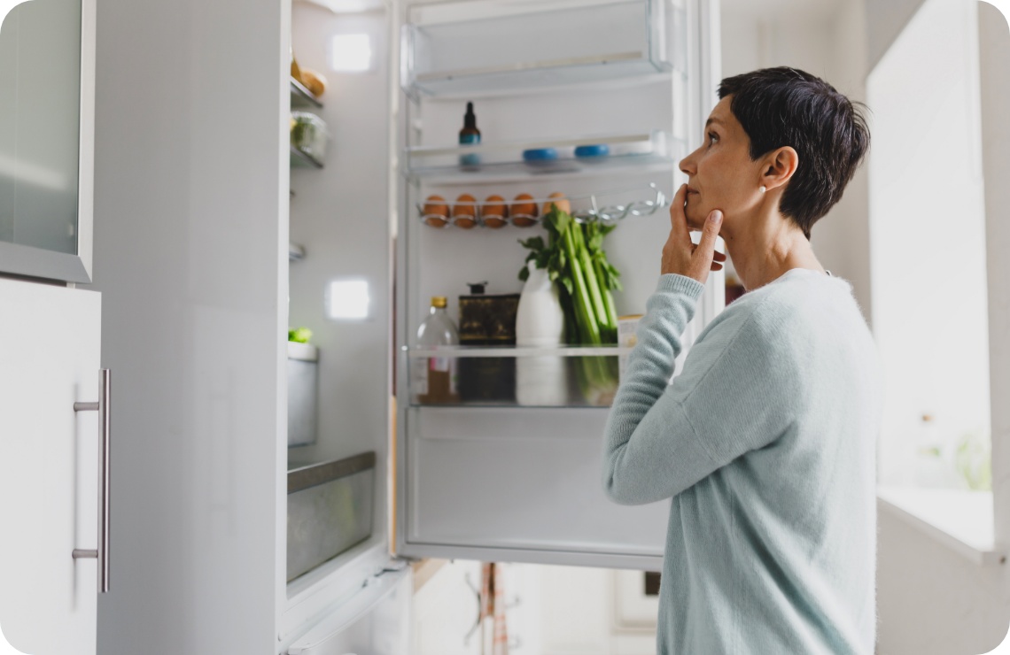 Frau in hellblauem Pullover schaut in einen offenem Kühlschrank gefüllt mit Eiern, Gemüse und Flaschen in einer hellen Küche.
