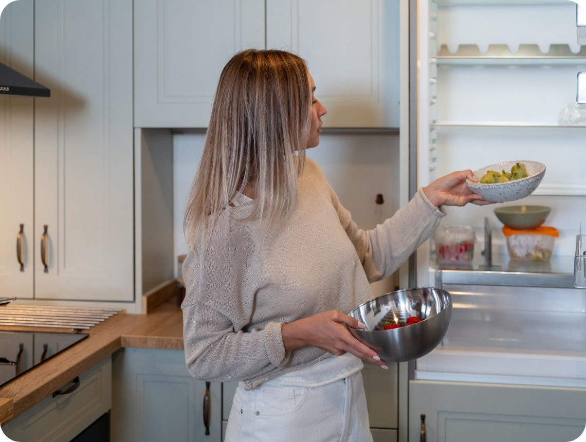Frau in heller Kleidung nimmt eine Schale mit Birnen aus einem fast leeren Kühlschrank und hält eine weitere Schale mit Erdbeeren in der Hand.