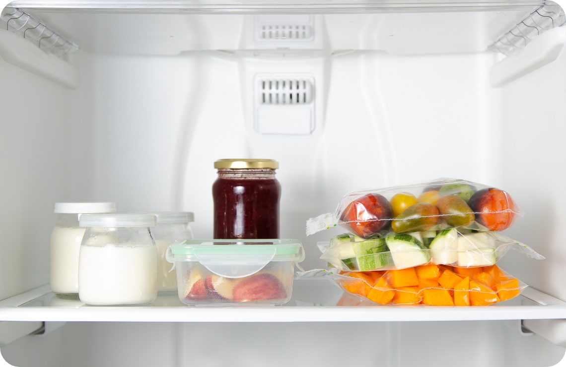 Kühlschrankfach mit Joghurtgläsern, Marmelade, geschnittenem Obst in einer Dose und verpacktem Gemüse wie Tomaten, Zucchini und Kürbis.