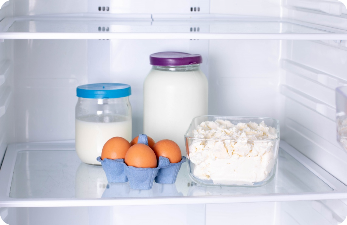 Kühlschrankfach mit Eiern in blauer Verpackung, zwei Milchgläsern und einer Glasschüssel mit frischem Hüttenkäse.