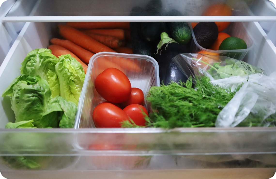 Blick in ein Gemüsefach gefüllt mit frischem Salat, Tomaten, Karotten, Gurken, Auberginen, Kräutern und Zitrusfrüchten.