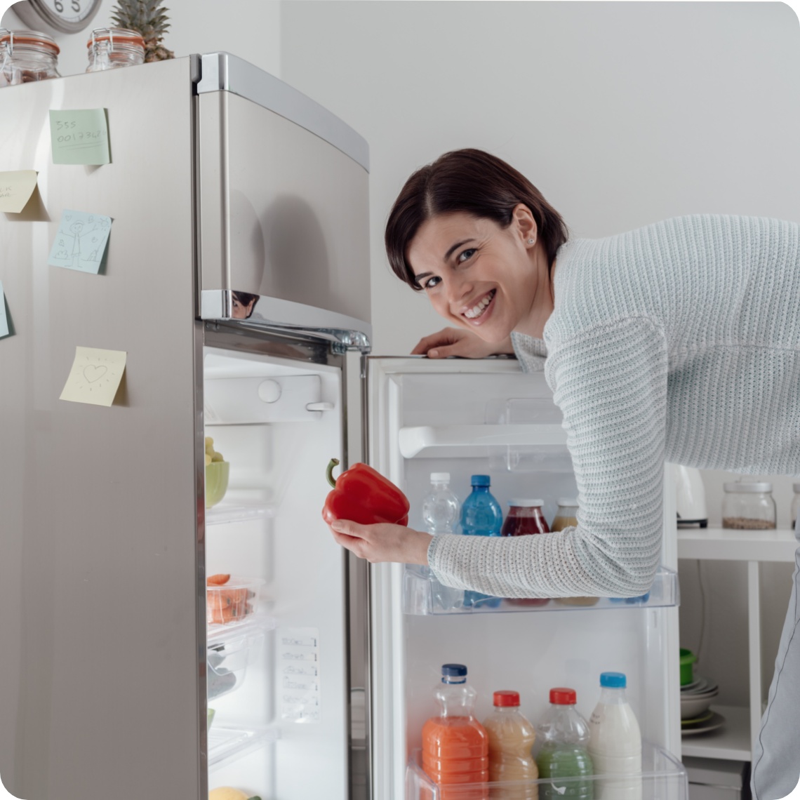 Lächelnde Frau holt eine rote Paprika aus einem offenen silbernen Kühlschrank.