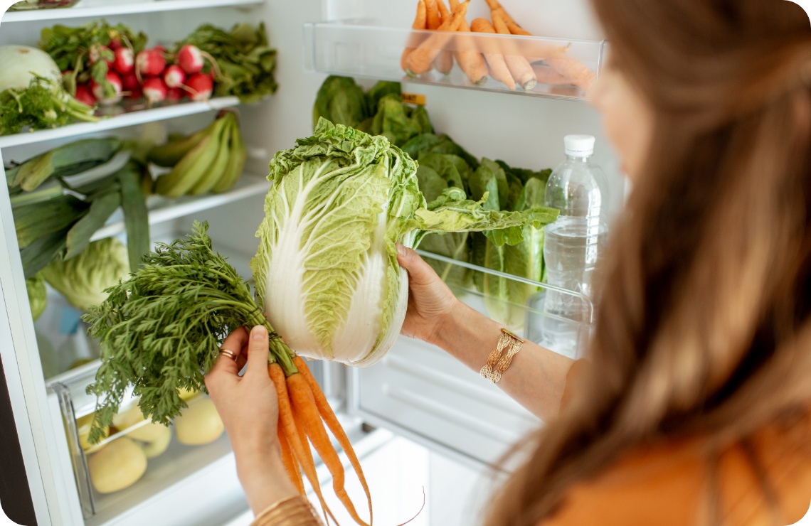Frau räumt frisches Gemüse, darunter Chinakohl und Karotten, in einen gut gefüllten Kühlschrank mit gesunden Lebensmitteln ein.