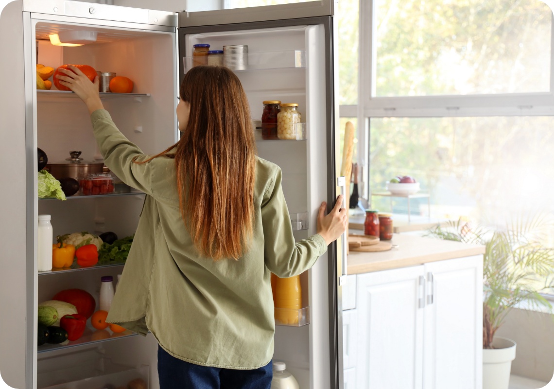 Frau mit langen Haaren räumt frische Paprika in einen gut gefüllten Kühlschrank in einer hellen, modernen Küche ein.