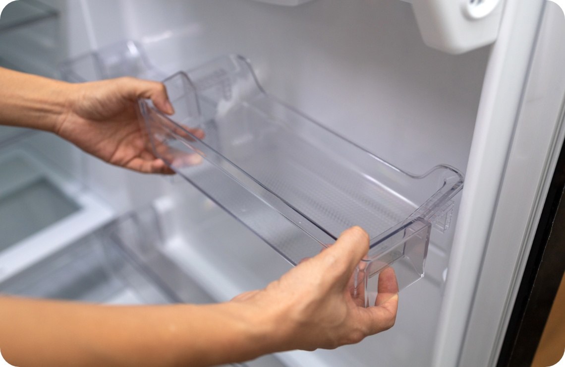 Nahaufnahme von Händen, die ein transparentes Kunststofffach in die Tür eines leeren Kühlschranks einsetzen oder es zur Reinigung entfernen.