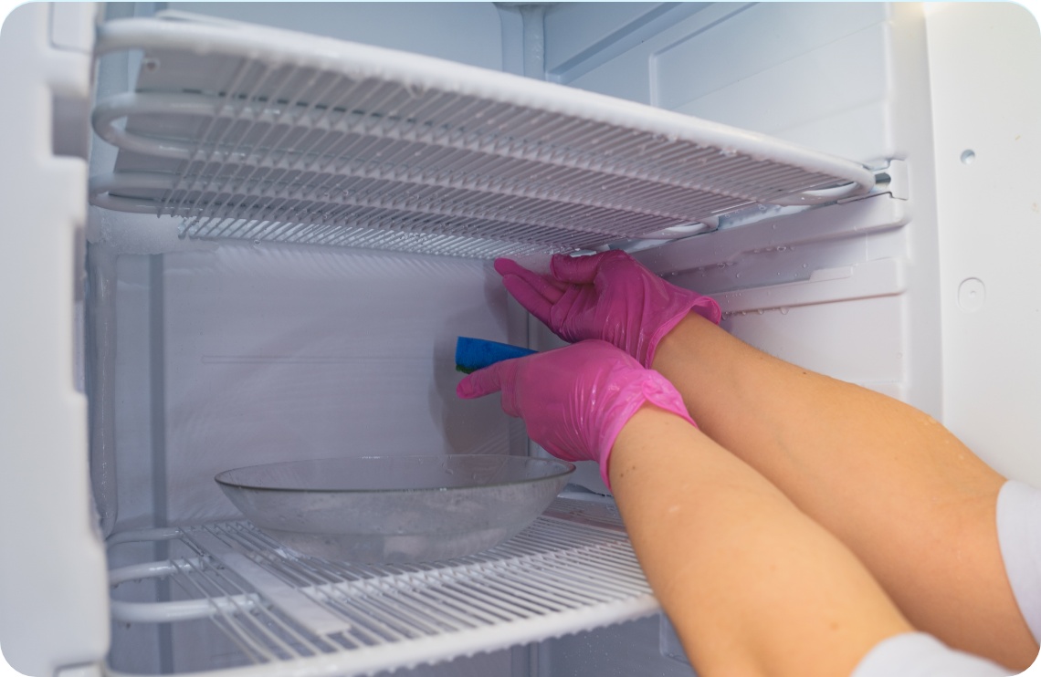 Person mit pinken Gummihandschuhen entfernt Eis aus einem vereisten Gefrierschrank mit einem blauen Schwamm.