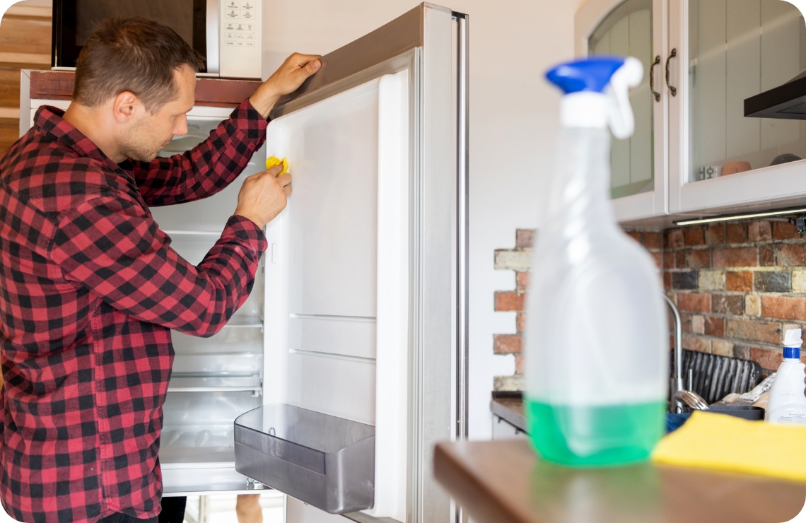 Mann in kariertem Hemd reinigt die Türdichtung eines offenen Kühlschranks mit einem gelben Tuch in einer rustikalen Küche.