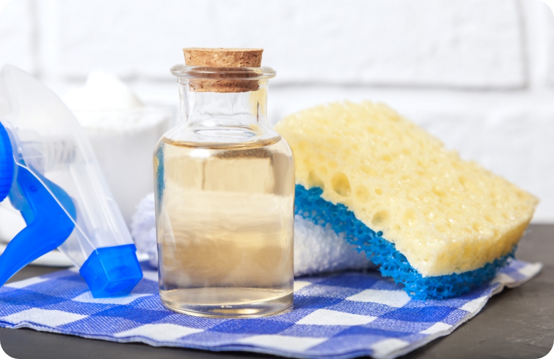 Glasflasche mit Essig, gelber Schwamm, Sprühflasche und Natron auf blau-weiß kariertem Tuch.