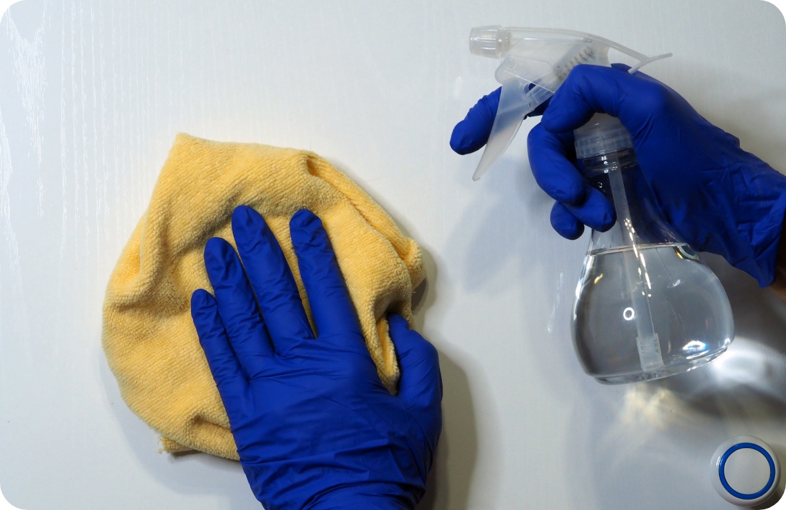 Person mit blauen Handschuhen reinigt eine Oberfläche mit gelbem Mikrofasertuch und transparenter Sprühflasche.