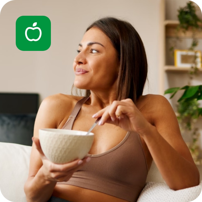 Frau in Sportkleidung sitzt auf einem weißen Sofa und isst lächelnd eine Bowl.