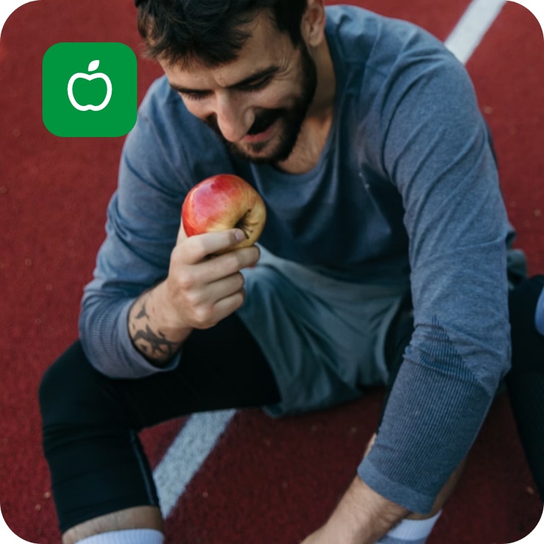 Zwei Personen in Sportkleidung sitzen auf einer Laufbahn und essen lächelnd Äpfel nach einem Training.