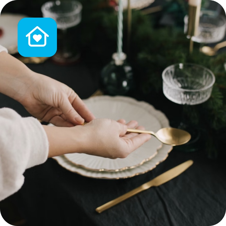 Hände halten einen goldenen Löffel vor einem, mit Tannenzweigen und weißem Geschirr, gedeckten Tisch.
