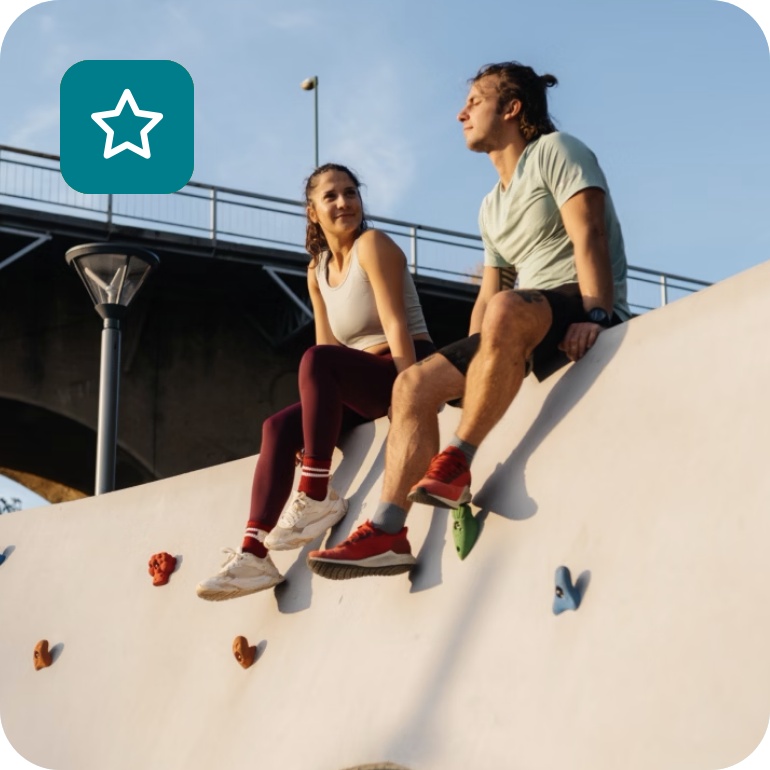 Zwei junge Erwachsene sitzen auf einer Kletterwand im Freien, lächeln und genießen das Sonnenlicht, mit Brücke im Hintergrund.