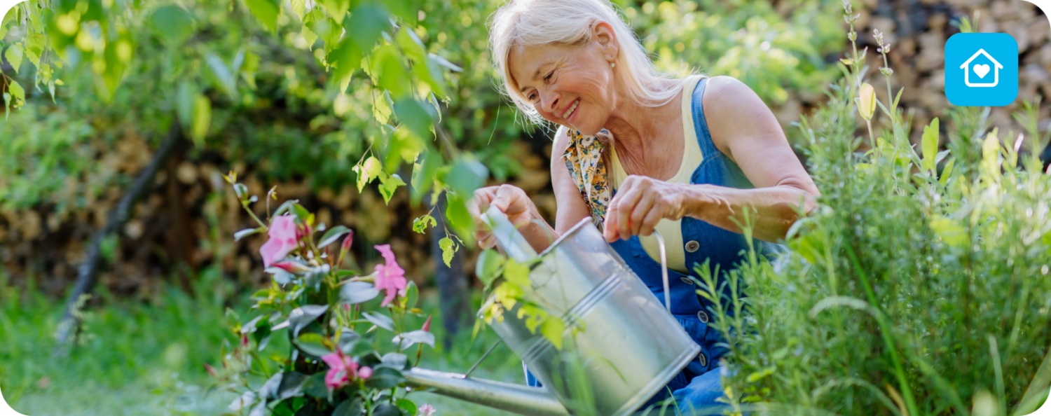 Ältere Frau mit blonden Haaren gießt mit einer Metallkanne rosa Blumen in einem grünen Garten, umgeben von Pflanzen.