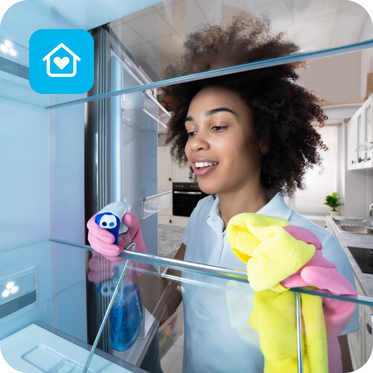 Frau mit Handschuhen reinigt einen leeren Kühlschrank mit Sprühreiniger und gelbem Mikrofasertuch in moderner Küche.