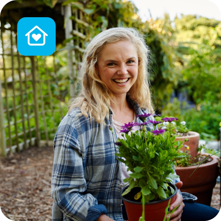 Eine Frau in einem grünen Garten mit einer blühenden Topfpflanze in der Hand.