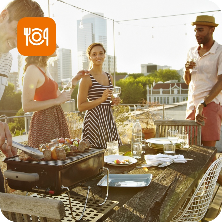 Gruppe junger Leute grillt und trinkt gemeinsam bei Sonnenuntergang auf städtischer Dachterrasse.