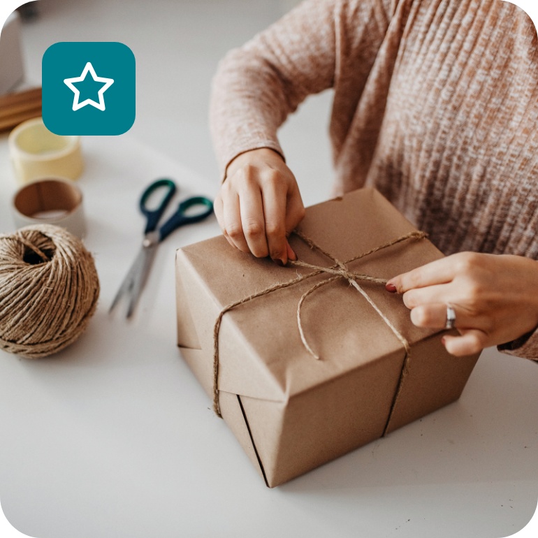 Frau verpackt Geschenk in braunem Papier mit Juteschnur, umgeben von Verpackungsmaterialien.