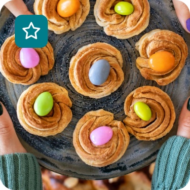 Teller mit Ostergebäck in Nestform, belegt mit bunten Schokoladeneiern, gehalten von Händen mit farbig lackierten Nägeln.