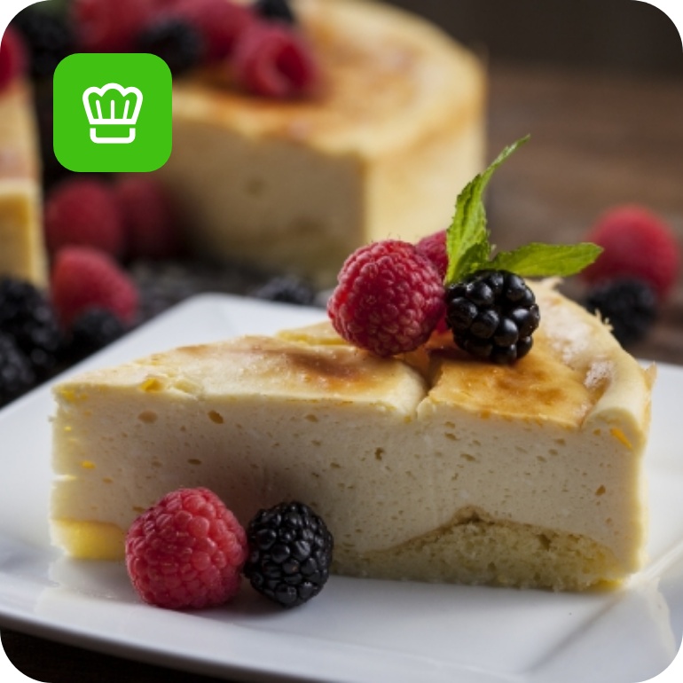 Stück Käsekuchen mit Himbeeren und Brombeeren auf weißem Teller, im Hintergrund ganze Torte.