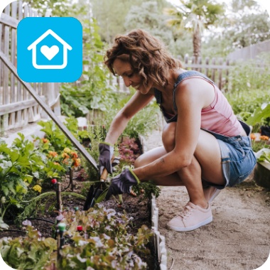 Frau hockt in kurzer Jeans-Latzhose, Tanktop und Gartenhandschuhen in einem mit Holzlatten eingezäunten Garten auf einem Weg. Links und rechts von ihr sind Beete angelegt, sie gräbt in dem linken Beet mit einer kleinen Schaufel.