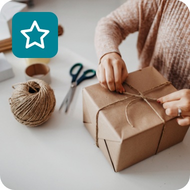 Frau verpackt Geschenk in braunem Papier mit Juteschnur, umgeben von Verpackungsmaterialien.