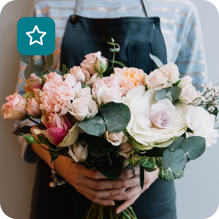 Floristin mit dunkelgrüner Schürze hält Blumenstrauß aus weißen und hellrosafarbenen Blumen.