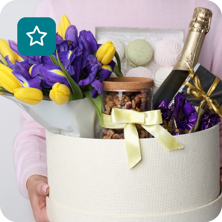 Eine junge Frau hält einen Geschenkkorb mit Sekt, Nüssen, Snacks und Blumen.