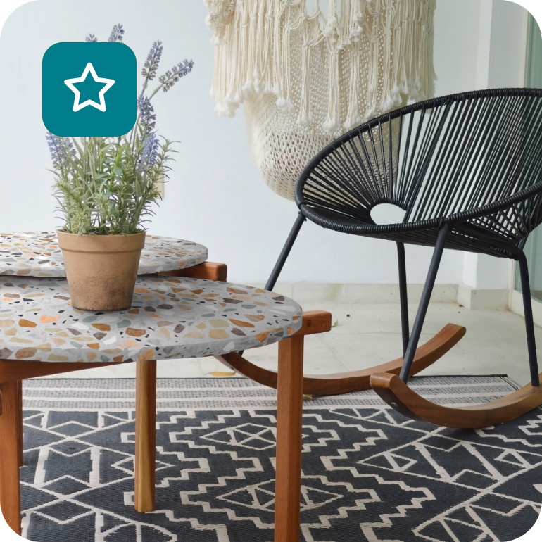 Zwei unterschiedlich große Tischplatten in Terrazzo-Optik mit Tischbeinen aus Holz zusammen mit runden Rattan-Schaukelstühle auf einem Balkon.