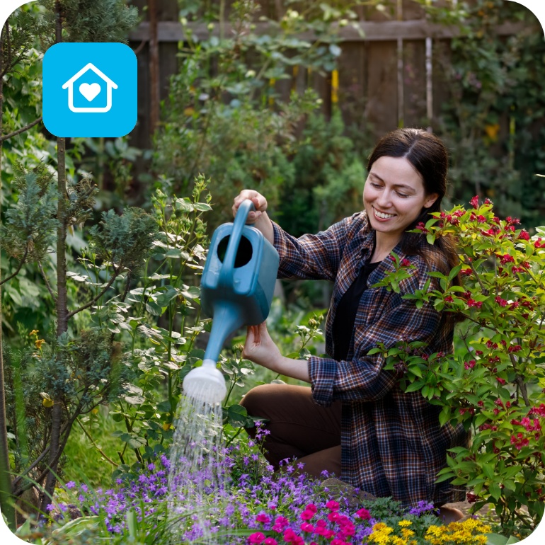 Frau gießt mit blauer Gießkanne bunte Blumen in grünem Garten, umgeben von Sträuchern und Blüten.