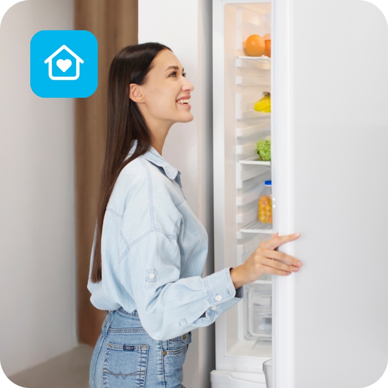 Junge Frau öffnet lächelnd einen weißen Kühlschrank in moderner Küche mit Obst und Gemüse im Inneren.